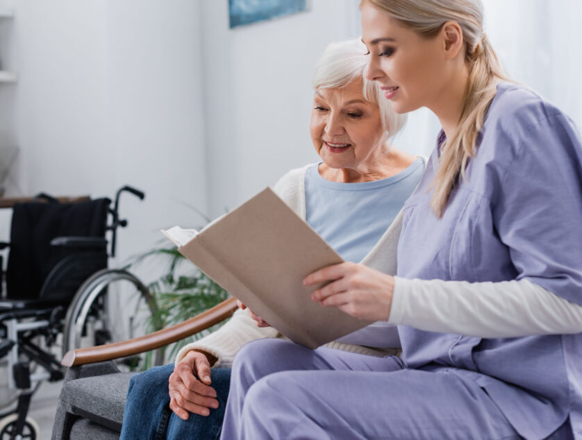 Young Social Worker Reading Book To Senior Woman While Sitting On Sofa young social worker reading book to senior woman while sitting on sofa